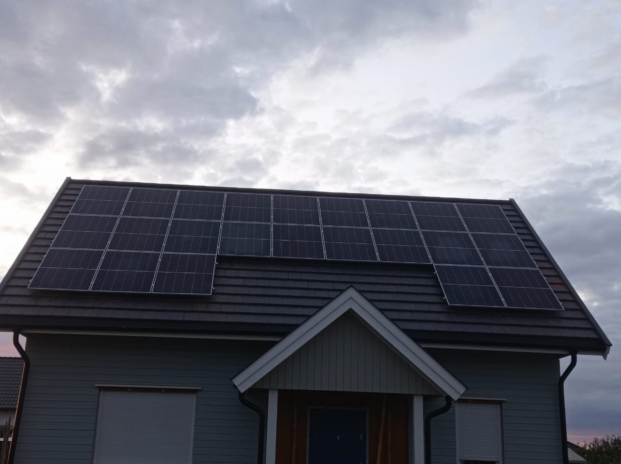 Panele słoneczne na dachu domu jednorodzinnego z szarą dachówką i elewacją. Pochmurne niebo w tle. Widok z dołu, lekki kąt.