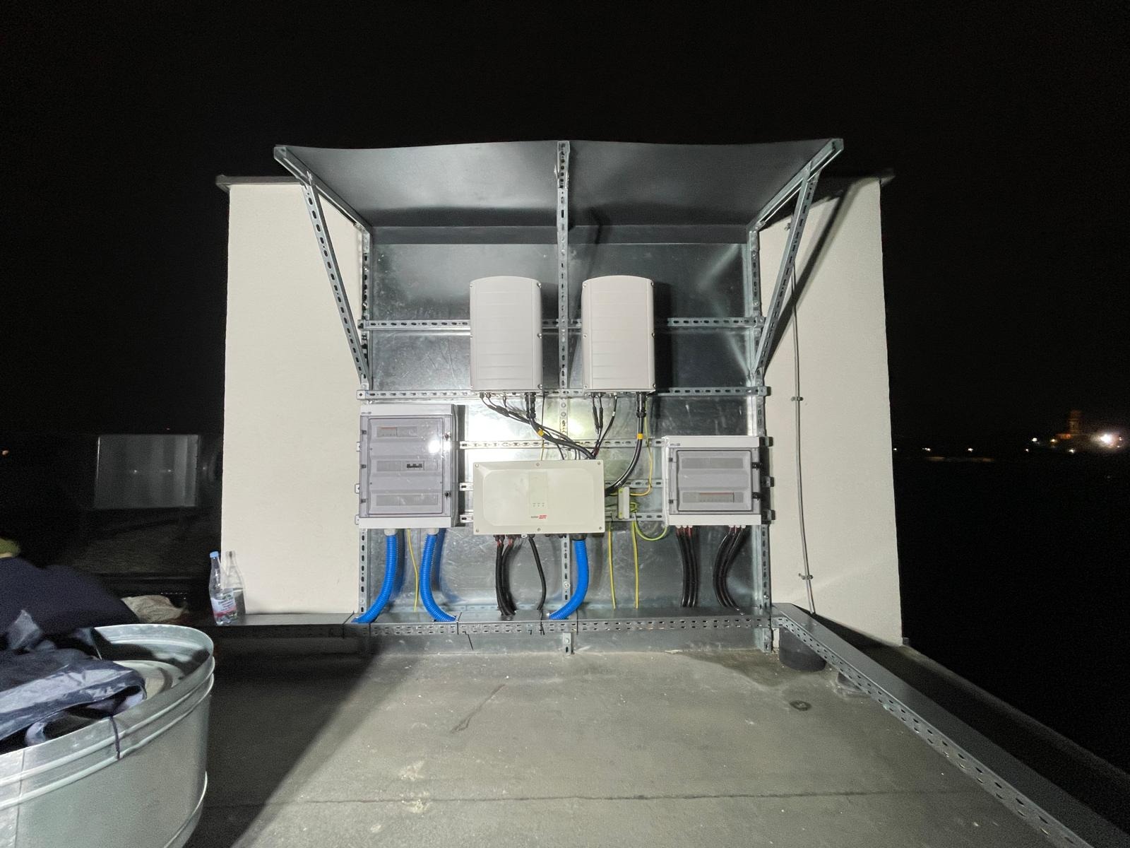 Instalacja elektryczna z osprzętem na dachu, zabezpieczona metalową konstrukcją. Widoczne kable, skrzynki rozdzielcze i urządzenia. Nocne ujęcie.