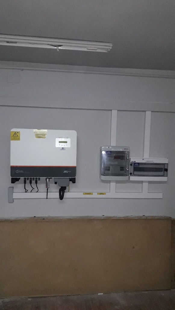 Zamontowany inwerter fotowoltaiczny Solis z okablowaniem i licznikiem na ścianie wewnątrz budynku. Widoczne elementy instalacji elektrycznej.