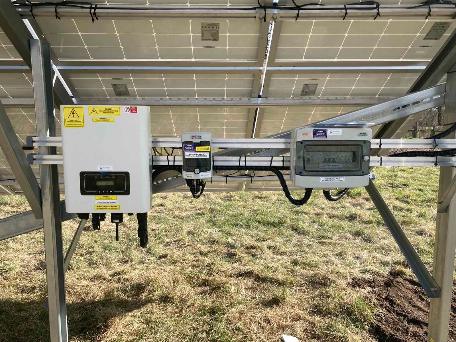 Instalacja paneli słonecznych z widocznym inwerterem, zabezpieczeniami i skrzynką przyłączeniową na tle trawy. Widok od spodu konstrukcji.