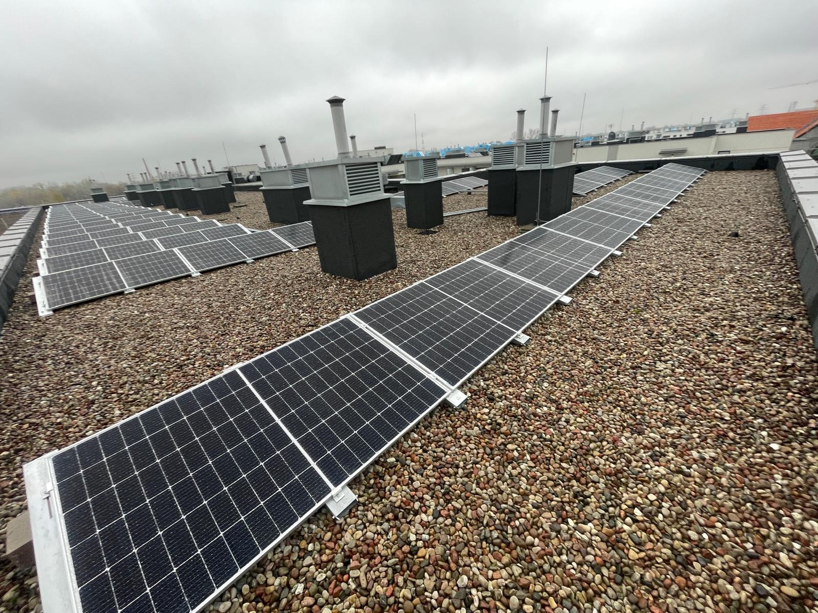 Rząd paneli słonecznych na żwirowym dachu budynku z wentylacją. Widok z góry, pochmurne niebo. Instalacja fotowoltaiczna na dachu przemysłowym.