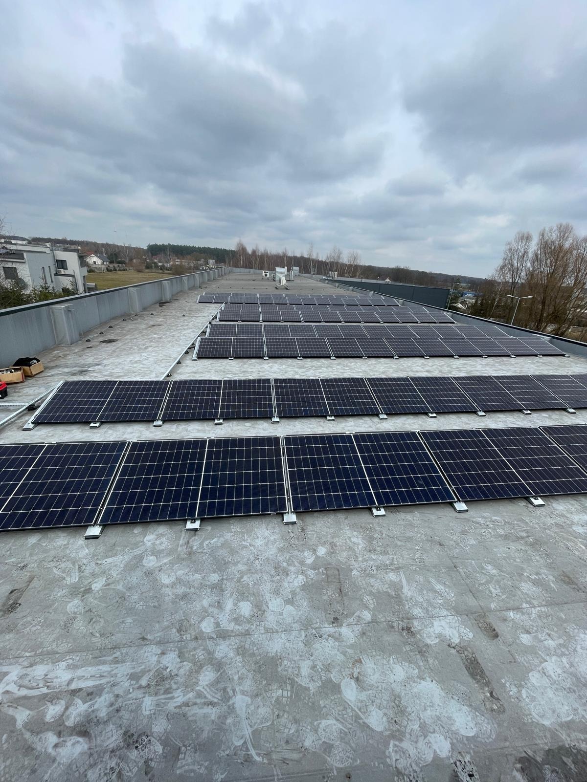 Rząd paneli słonecznych na płaskim dachu budynku, widok z góry. W tle zabudowa miejska i pochmurne niebo. Widoczne ślady na dachu.