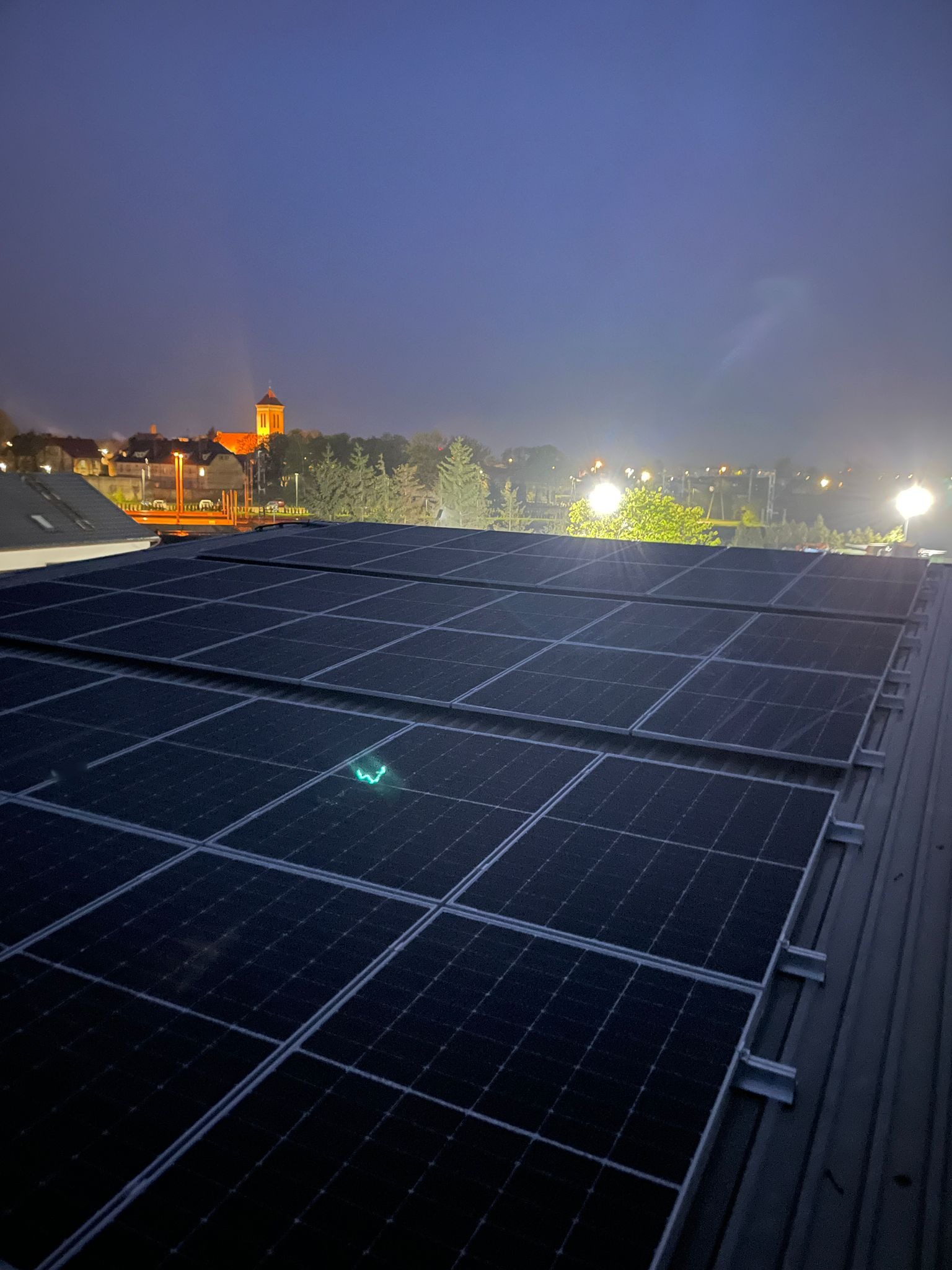 Panele słoneczne na dachu budynku o zmierzchu, w tle widoczny kościół i oświetlone budynki. Nocne ujęcie instalacji.