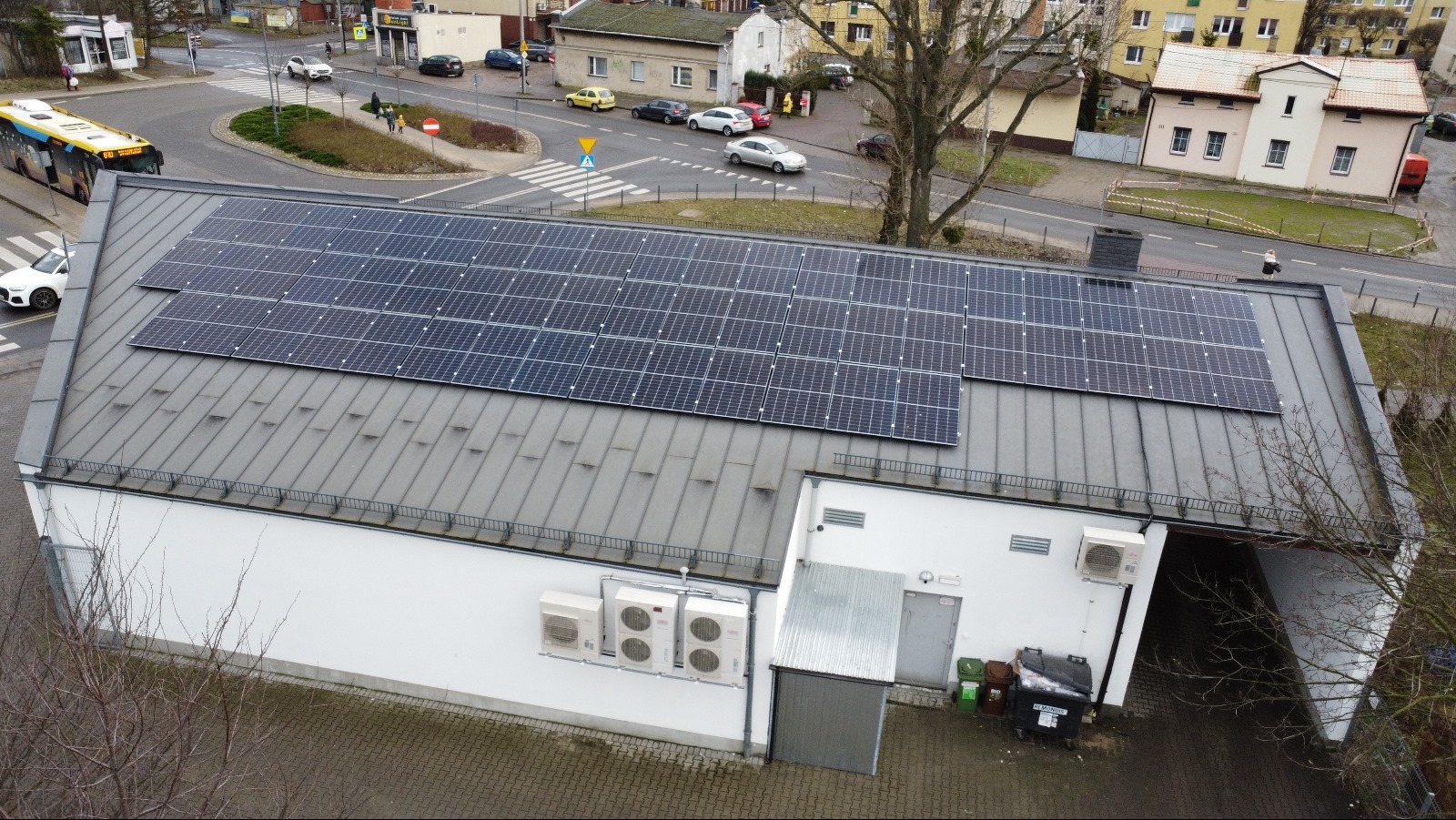 Instalacja paneli na dachu budynku z blachy trapezowej, widok z góry na skrzyżowanie i ruch uliczny w tle. Widoczne klimatyzatory na ścianie budynku.