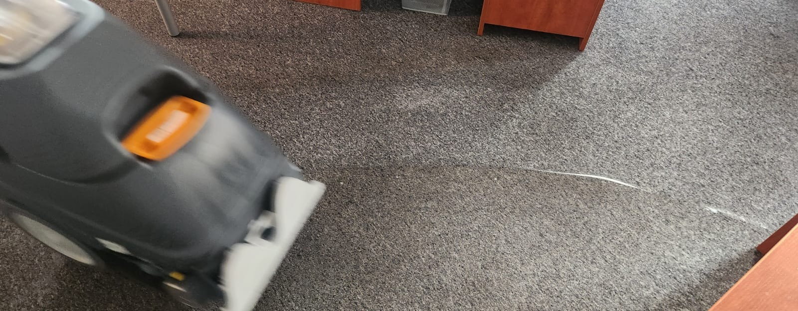 Szara wykładzina dywanowa czyszczona maszyną z widoczną smugą wilgoci po czyszczeniu. Fragmenty mebli biurowych w tle.