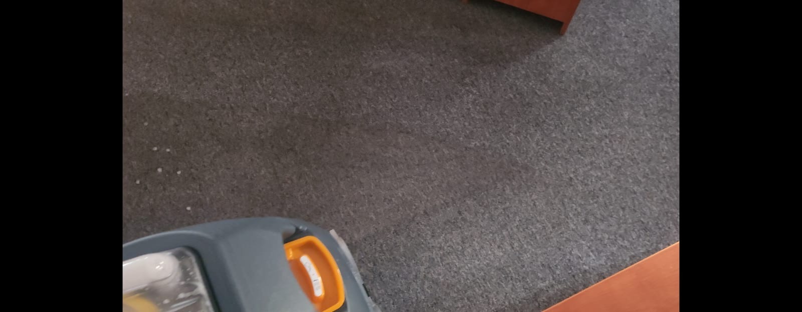 Szary dywan czyszczony maszyną z widocznymi plamami i fragmentem mebla. Ujęcie z góry, prezentujące proces prania dywanu.