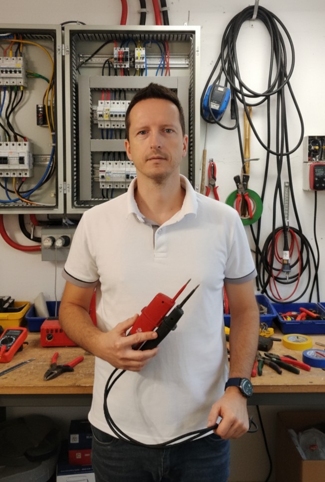 Elektryk prezentuje miernik napięcia na tle rozdzielnicy i narzędzi. Widoczne kable, bezpieczniki i osprzęt. Profesjonalny warsztat elektryczny.