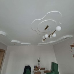 Pejta Decon - Dekoracyjny sufit podwieszany z oświetleniem punktowym i ozdobnym żyrandolem w jasnym wnętrzu. Na ścianie drewniana mapa świata.