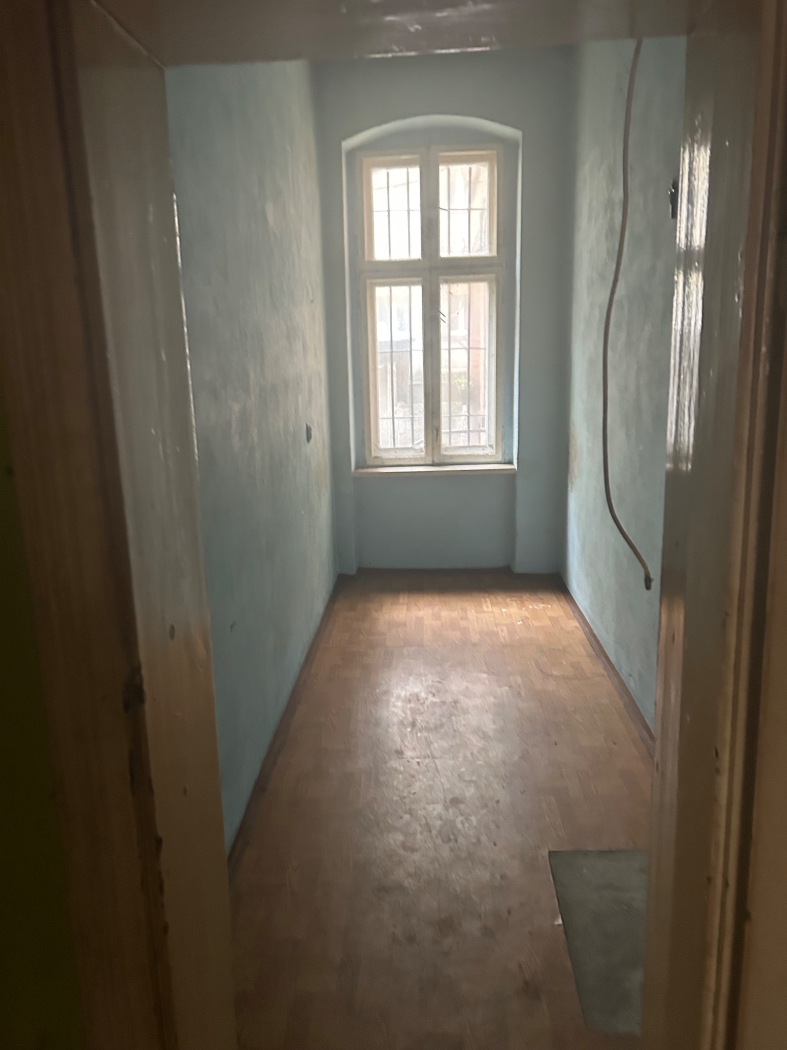Pusty pokój z drewnianą podłogą i błękitnymi ścianami, widok zza framugi drzwi. Okno z kratami w tle. Widoczne ślady użytkowania i przygotowania do prac remontowych.