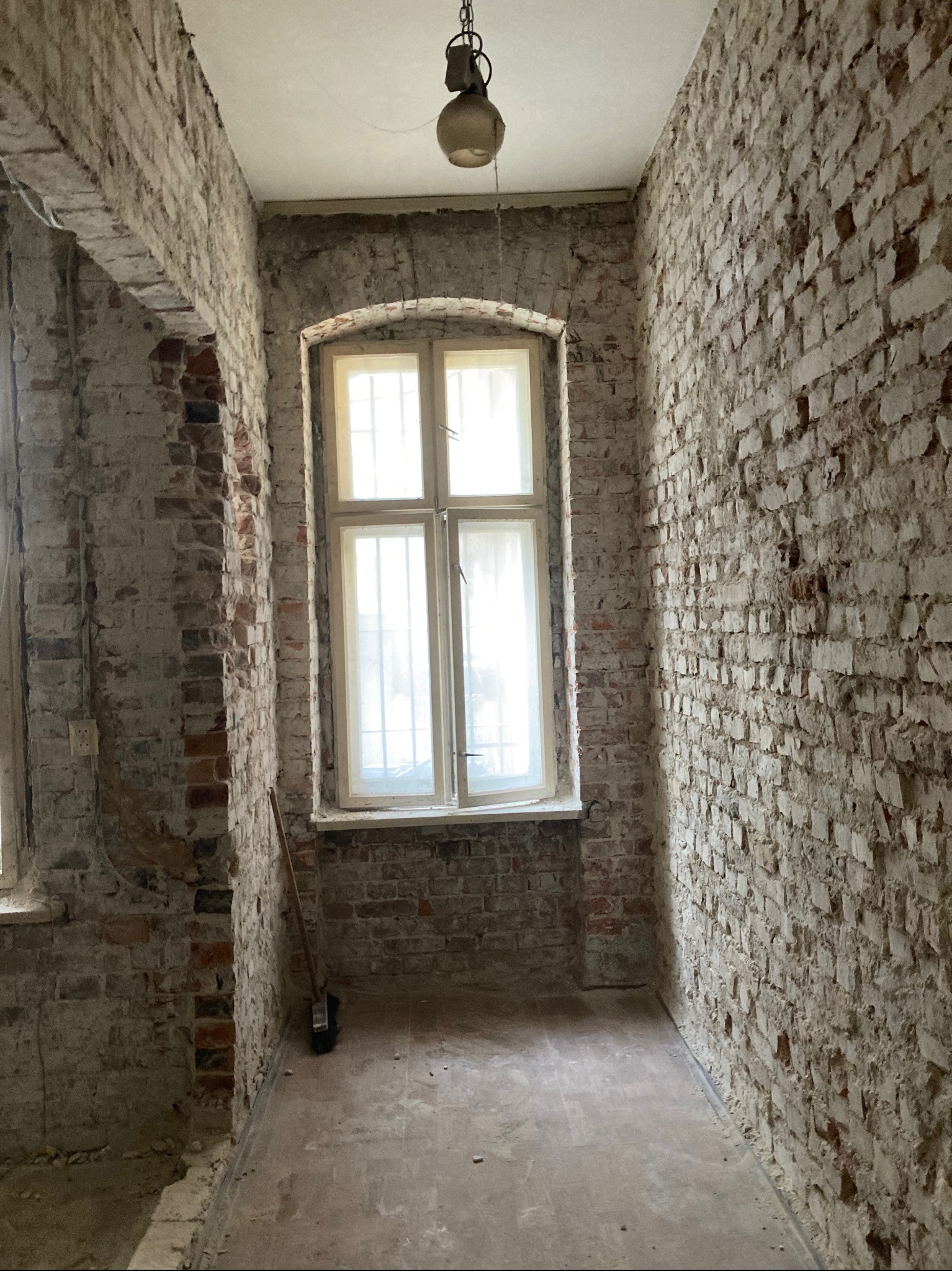 Wnętrze w trakcie renowacji: surowe ceglane ściany z oknem, widoczne ślady po usuniętych tynkach, podłoga przygotowana do dalszych prac. Wisząca lampa dodaje klimatu.