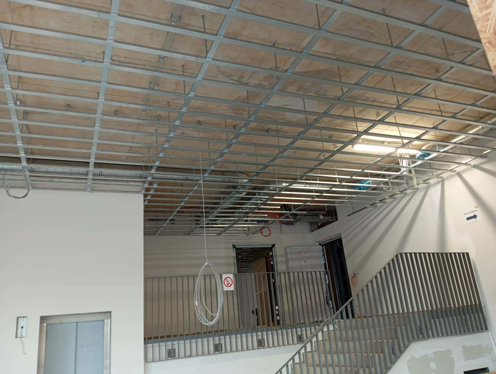 Szkielet sufitu podwieszanego z metalowych profili w jasnym wnętrzu budynku, widok z dołu na konstrukcję przed montażem płyt. Widoczne instalacje i przewody.