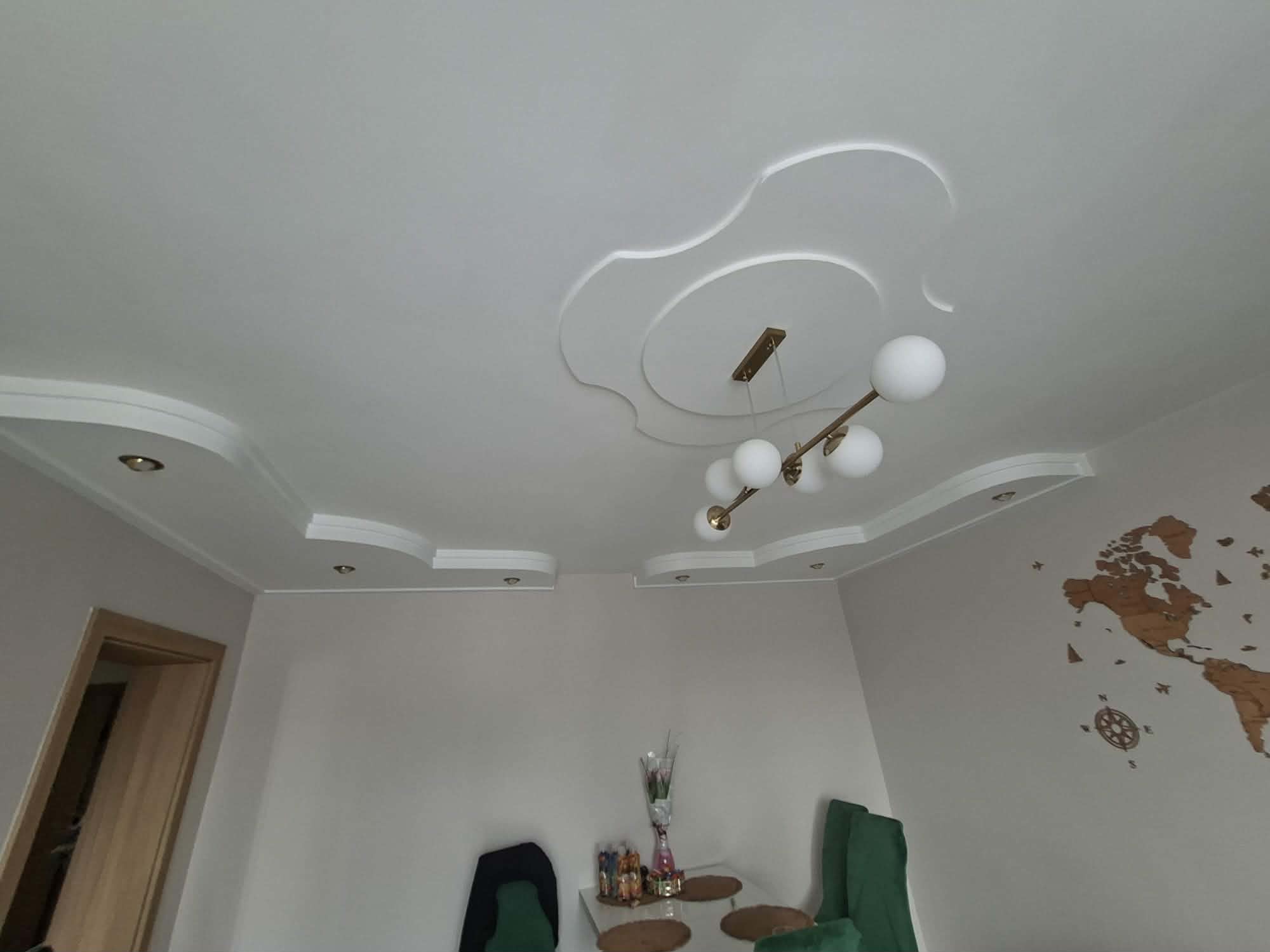 Dekoracyjny sufit podwieszany z oświetleniem punktowym i ozdobnym żyrandolem w jasnym wnętrzu. Na ścianie drewniana mapa świata.