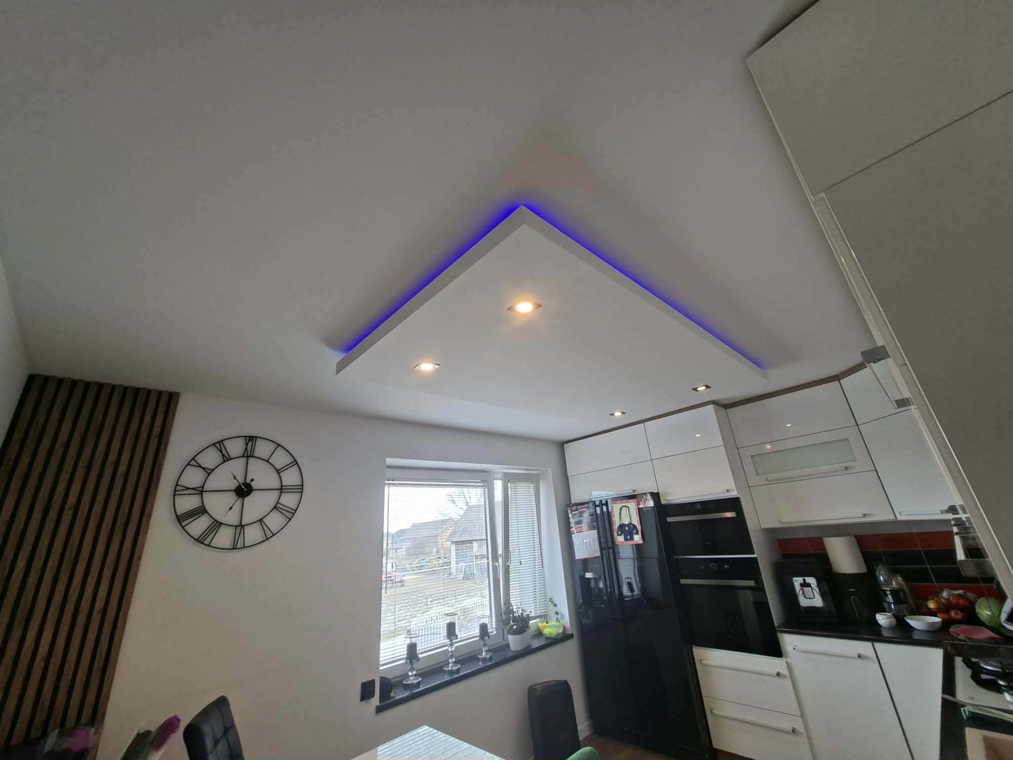 Nowoczesna kuchnia z podwieszanym sufitem w kształcie trójkąta, oświetlonym niebieskim światłem LED. Widoczny zegar na ścianie i zabudowa kuchenna w bieli.