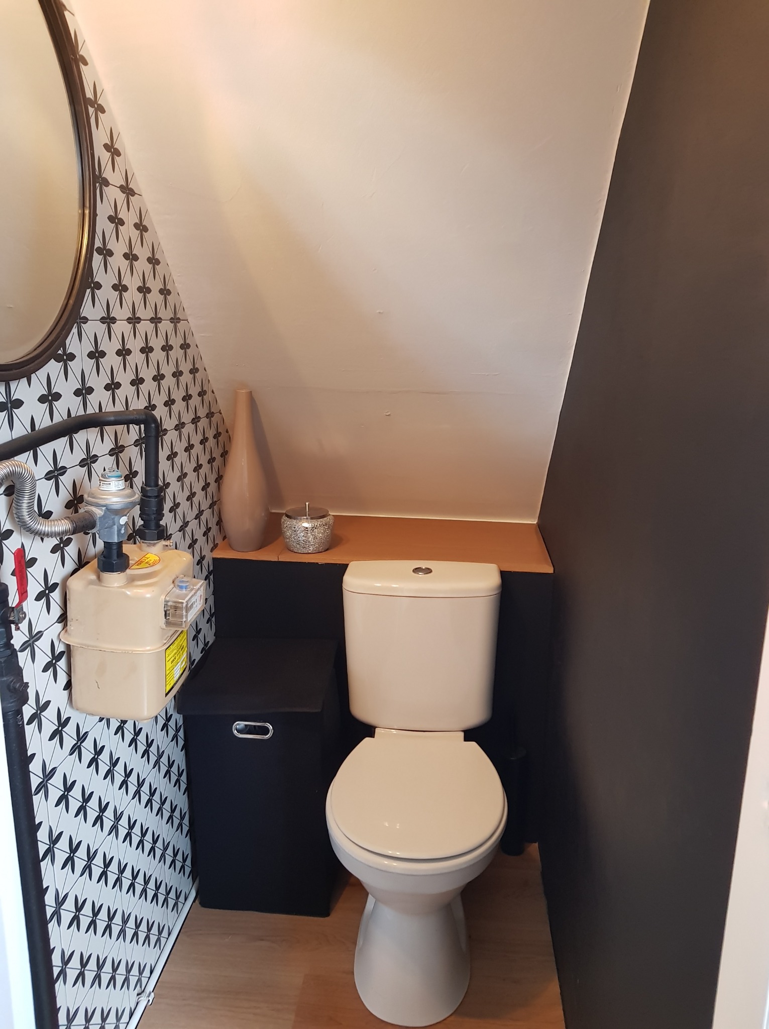 Kompaktowa toaleta w skosie poddasza: biała ceramika, wzorzyste płytki, czarne akcenty i drewniany blat. Miernik gazu widoczny na ścianie.