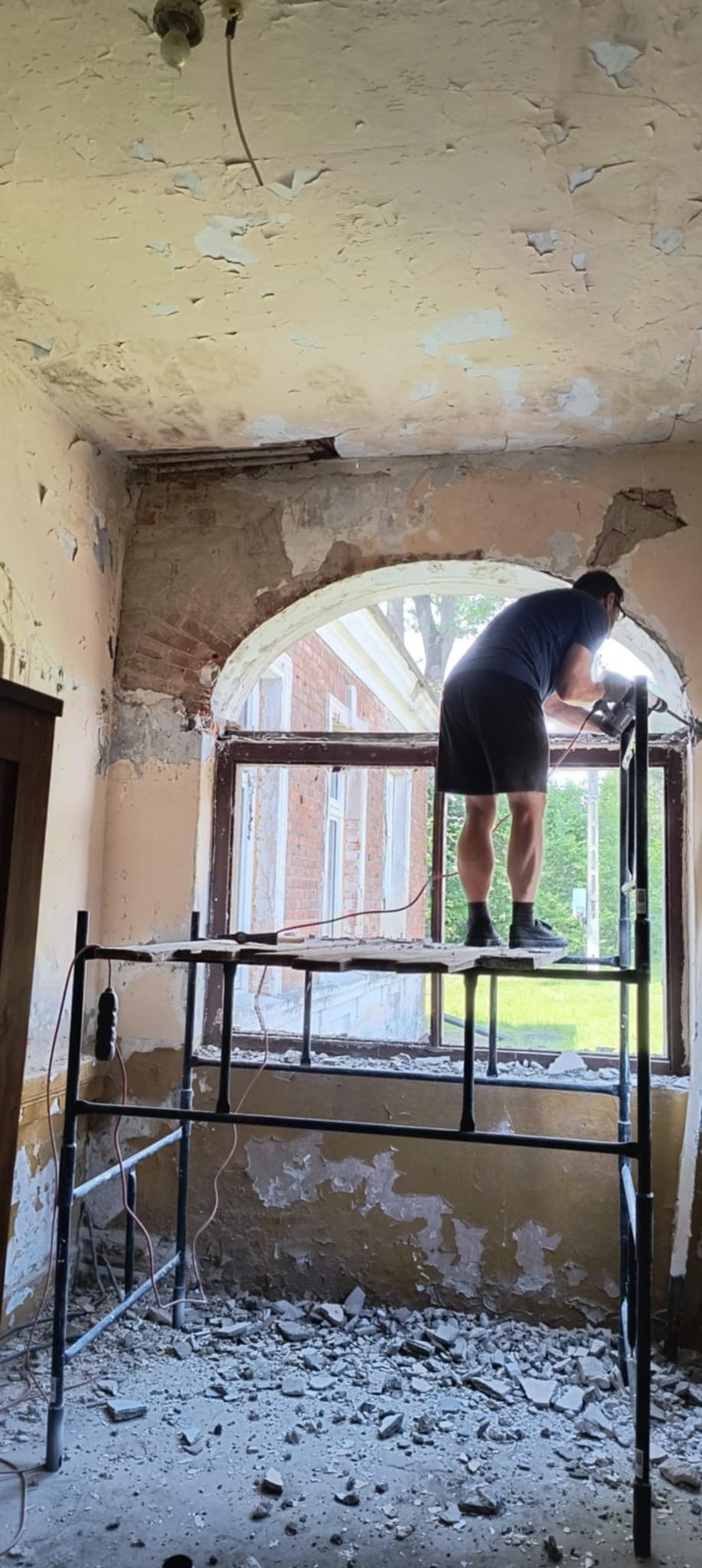 Pracownik na rusztowaniu, w trakcie skuwania tynku wokół okna z łukiem w starym budynku. Widoczne odsłonięte cegły i odpadający tynk.