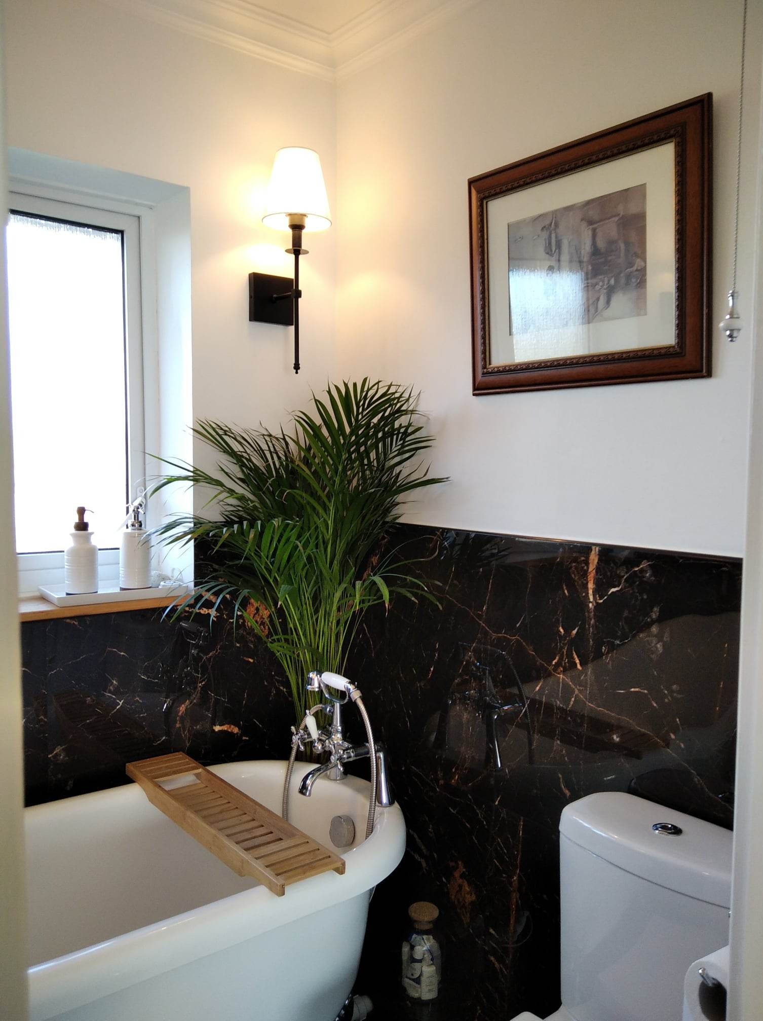 Elegancka łazienka z wanną na nóżkach, czarnymi płytkami imitującymi marmur i zieloną rośliną. Na ścianie kinkiet i obraz w drewnianej ramie.