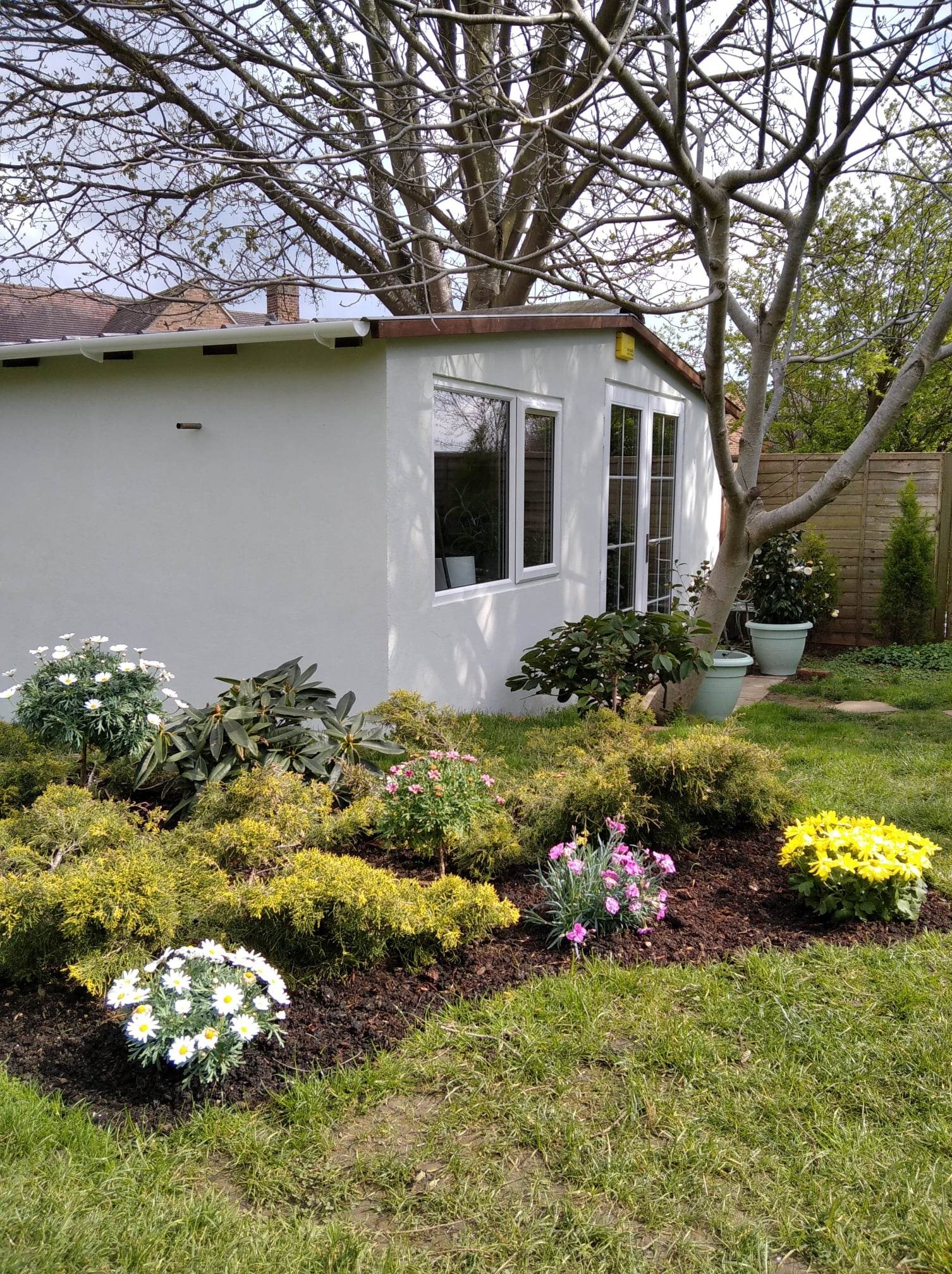 Otynkowany budynek gospodarczy z białymi oknami, otoczony rabatami kwiatowymi i drzewami w słoneczny dzień. Widoczne świeże nasadzenia i zadbany ogród.