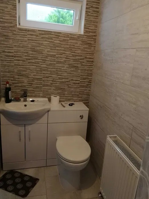 Jasna łazienka po remoncie: beżowe płytki imitujące kamień na ścianach, biała umywalka z szafką i toaleta kompaktowa. Małe okno z widokiem na zieleń. Grzejnik na ścianie.