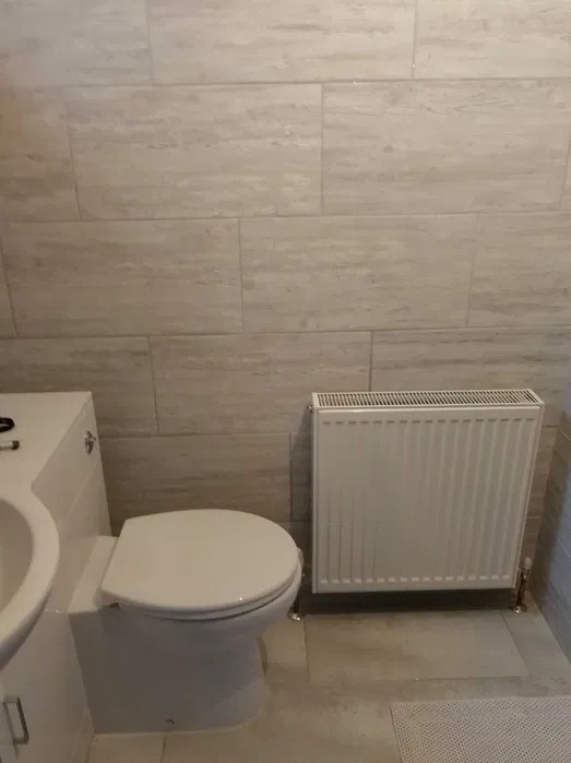 Jasna łazienka po remoncie: beżowe płytki imitujące drewno na ścianach, biała toaleta z umywalką i grzejnik. Minimalistyczny design, czyste linie.