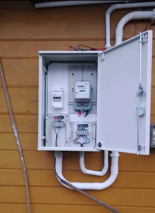 Otwarta skrzynka elektryczna z licznikiem i zabezpieczeniami na drewnianej ścianie. Widoczne białe rury instalacyjne i przewody. Ujęcie z bliska.