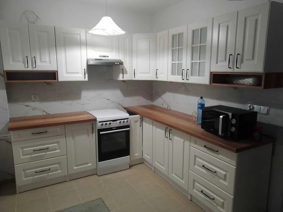 Jasna kuchnia z białymi szafkami i drewnianymi blatami. Widoczny sprzęt AGD: kuchenka i mikrofalówka. Marmurowe wykończenie ściany nad blatem. Nowoczesne oświetlenie.