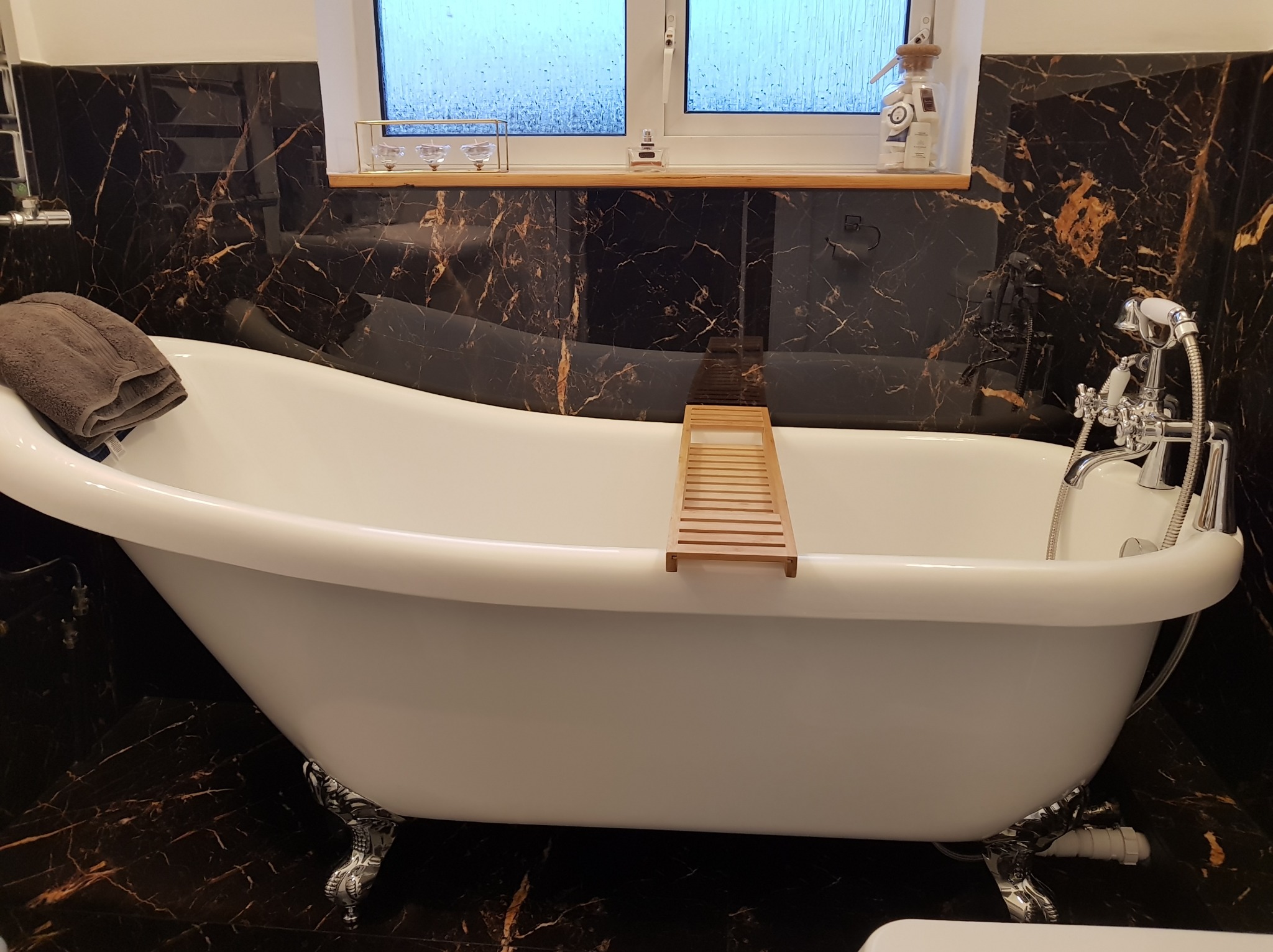 Elegancka, wolnostojąca wanna na ozdobnych nóżkach, z drewnianą półką i ręcznikiem, na tle czarnych płytek z marmurowym wzorem. Widok na okno.