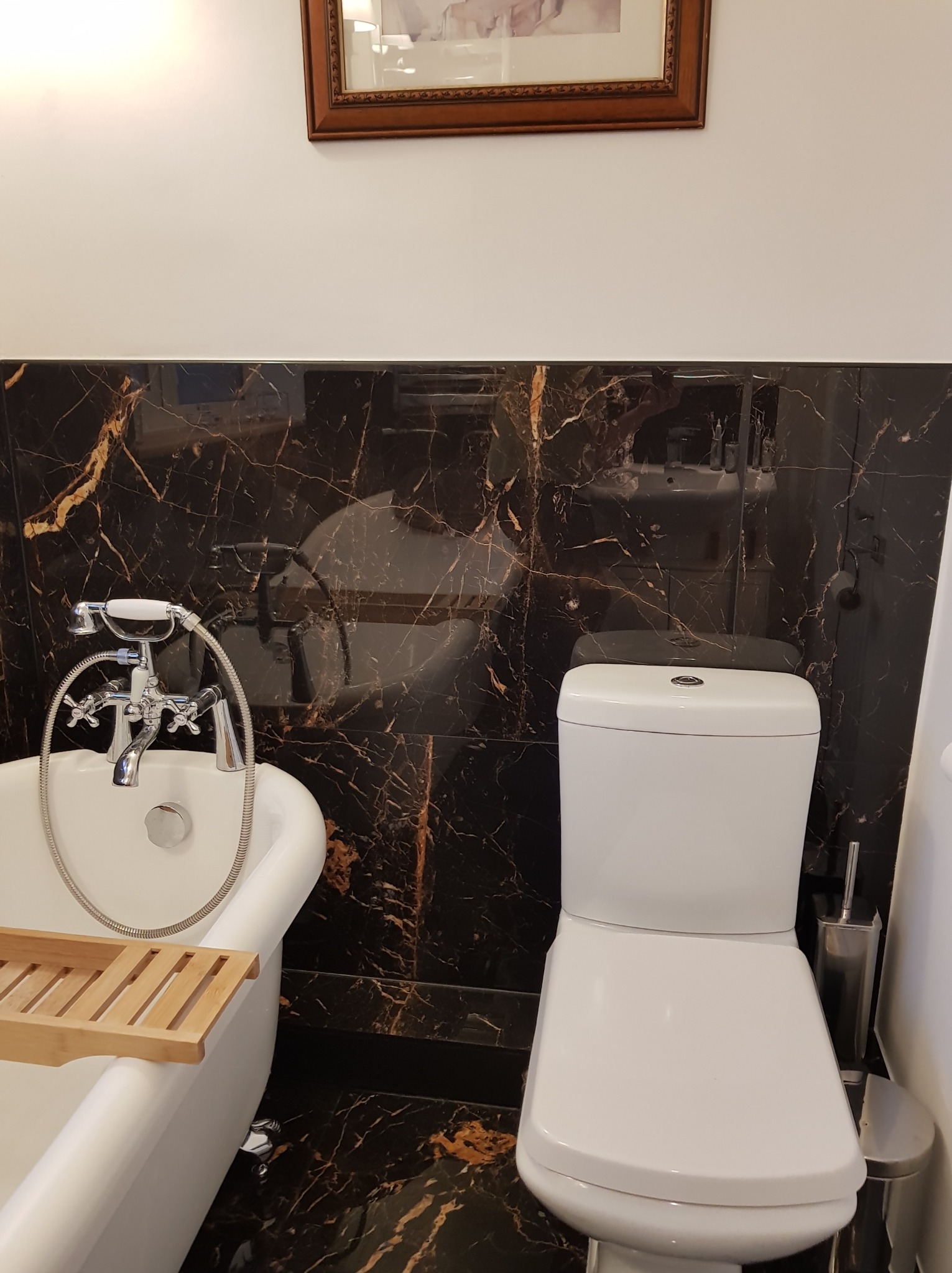 Elegancka łazienka z wanną na nóżkach i toaletą, wykończona czarnym marmurem z złotymi żyłami. Nad wanną drewniana półka, na ścianie obraz w ramie.