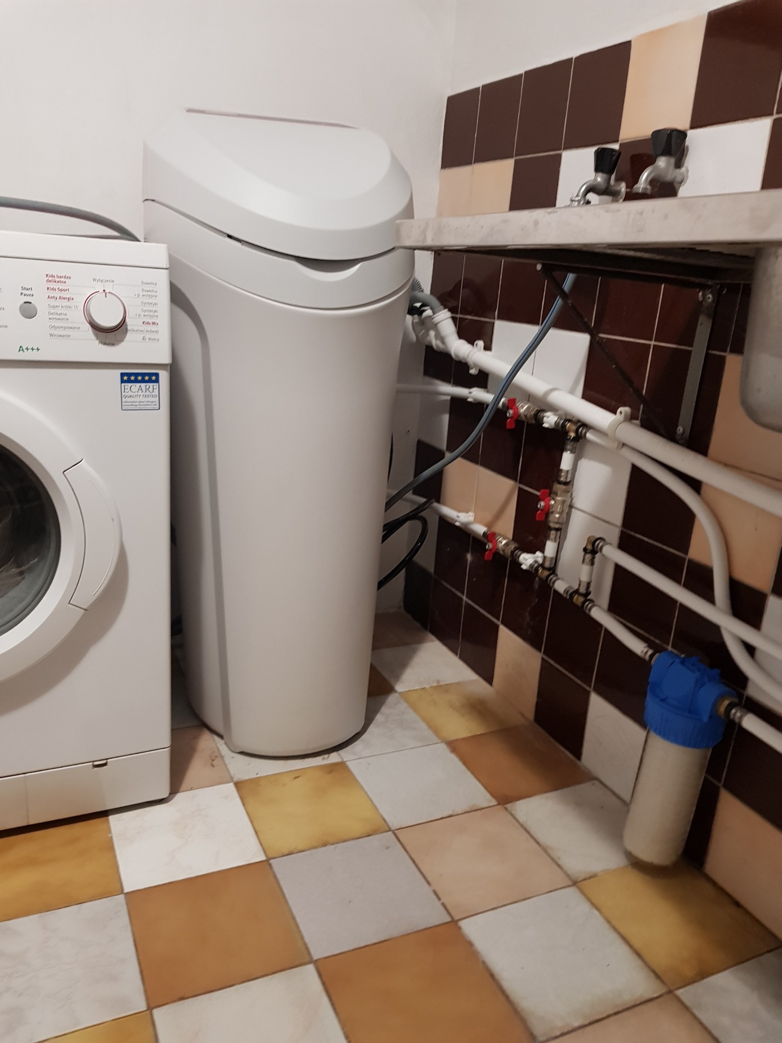 Biały zmiękczacz wody obok pralki w łazience z brązowo-beżowymi płytkami. Widoczne rury i zawory pod umywalką. Filtr wody na podłodze.