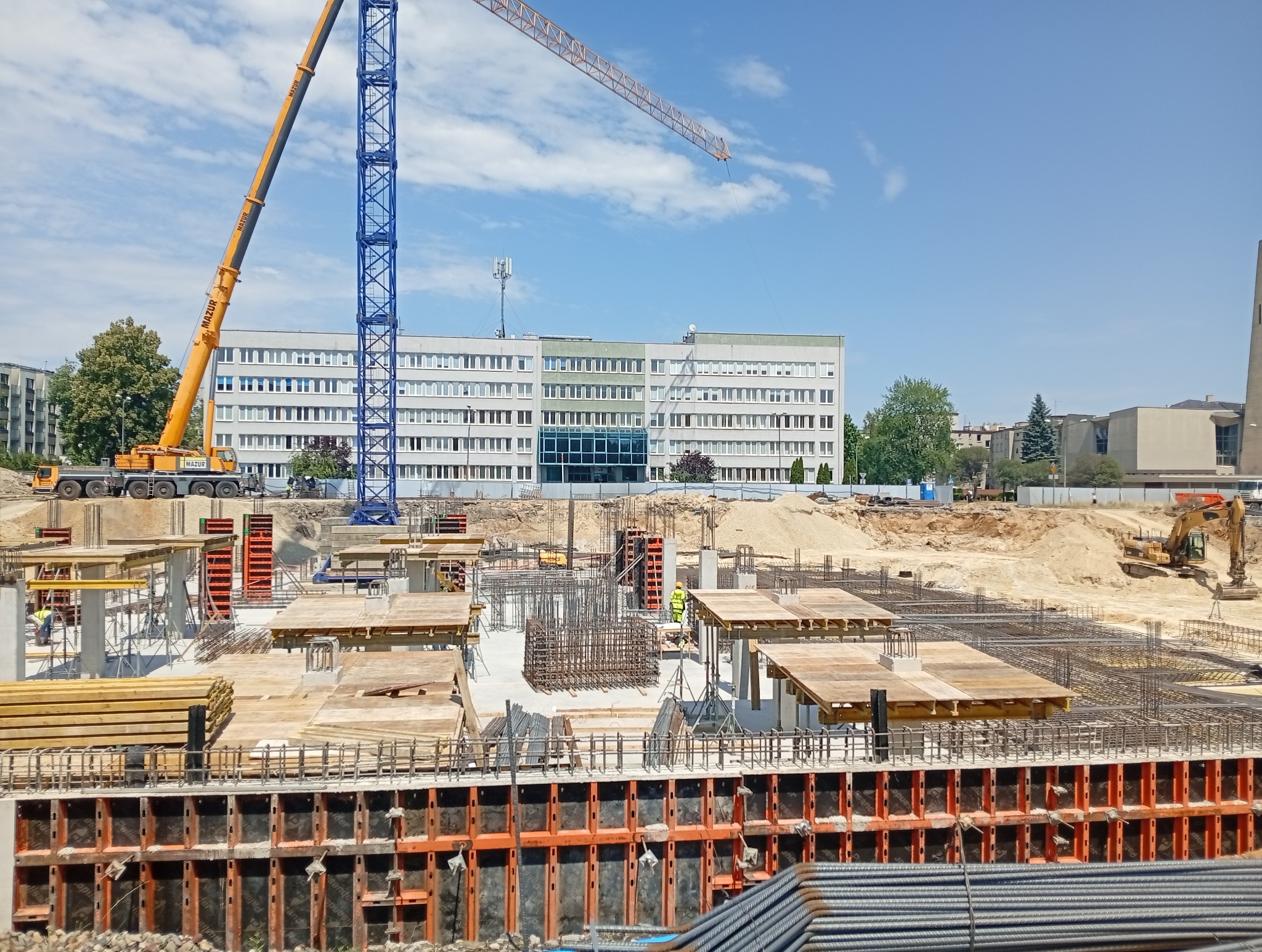 Szeroki kadr na plac budowy z widocznymi zbrojeniami, szalunkami, dźwigami i budynkiem w tle, w słoneczny dzień. Widoczny etap prac fundamentowych.