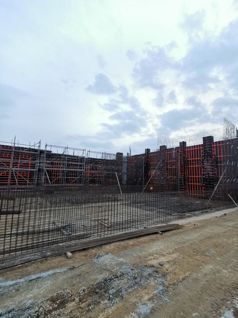 Siatka zbrojeniowa na budowie, widok z poziomu gruntu, szalunki w tle, pochmurne niebo. Elementy konstrukcyjne przygotowane do zalania betonem.