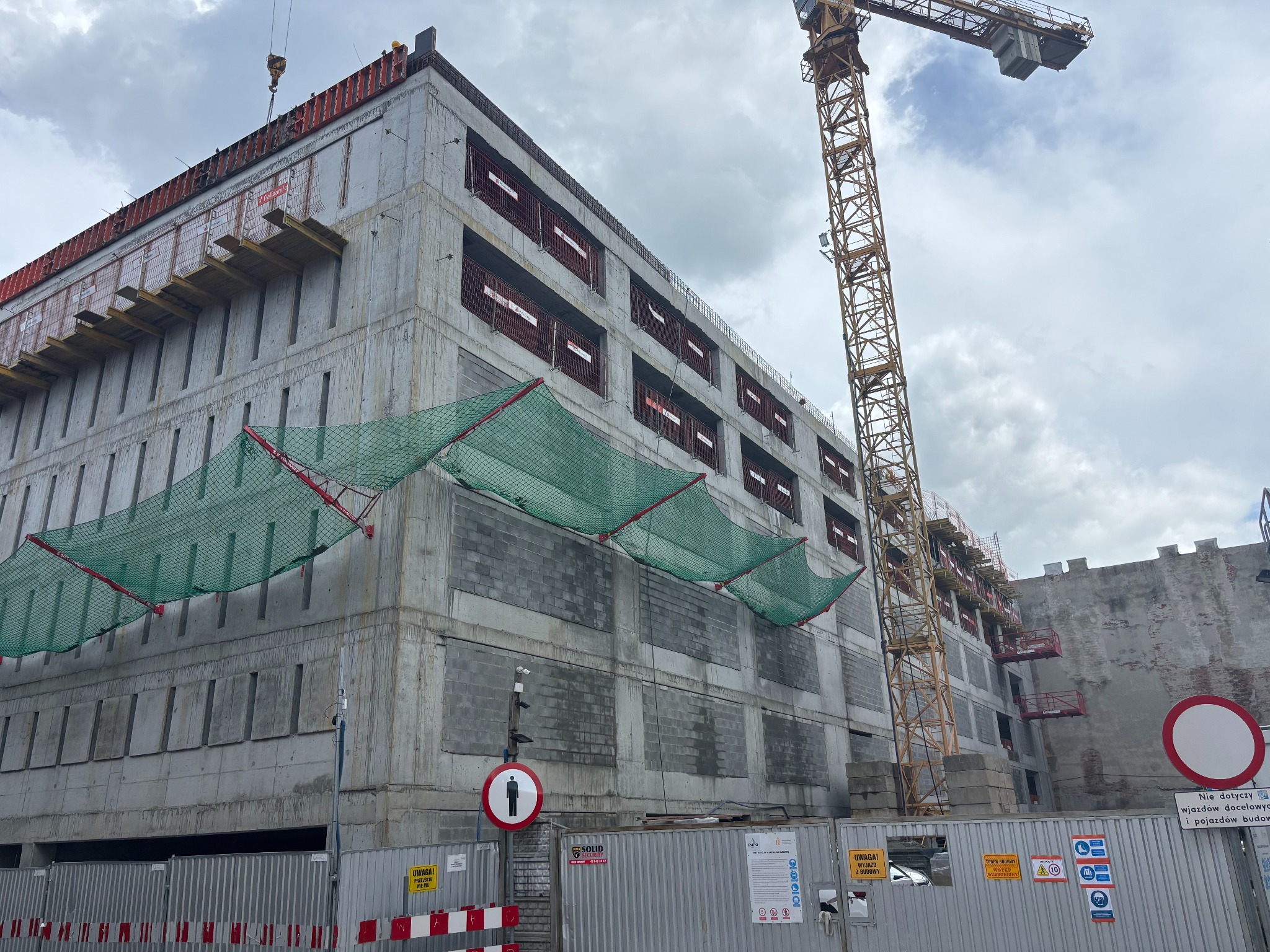 Budowa wielopiętrowego budynku z betonu, widoczny żuraw budowlany i zabezpieczenia siatką. Tablice informacyjne i ogrodzenie placu budowy.