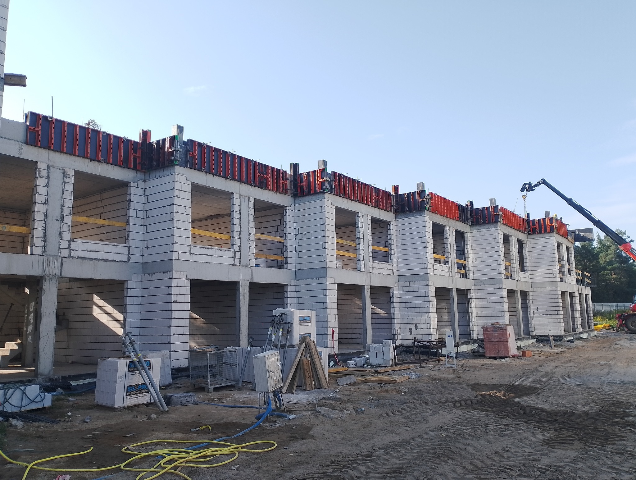 Budowa bliźniaka z szarej cegły silikatowej, widoczne szalunki stropowe w kolorze czerwonym, na placu budowy stoi żuraw.