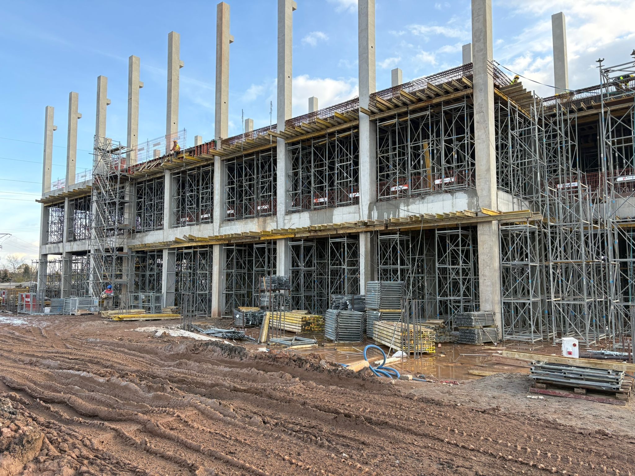 Wielopiętrowa konstrukcja w trakcie budowy z widocznymi zbrojeniami, szalunkami i betonowymi słupami. Na pierwszym planie błotniste podłoże z wyraźnymi śladami opon.