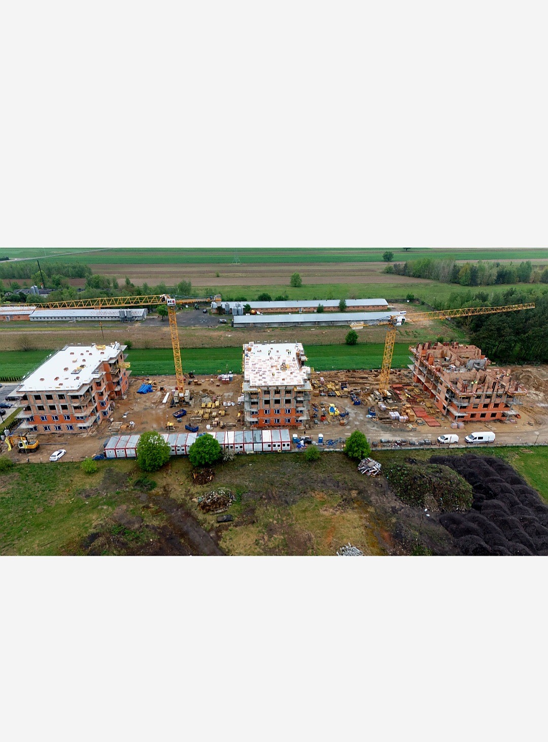 Budowa trzech budynków mieszkalnych w Warszawie, widok z drona. Na placu budowy kontenery, dźwigi i materiały budowlane. Jeden budynek w stanie surowym otwartym.