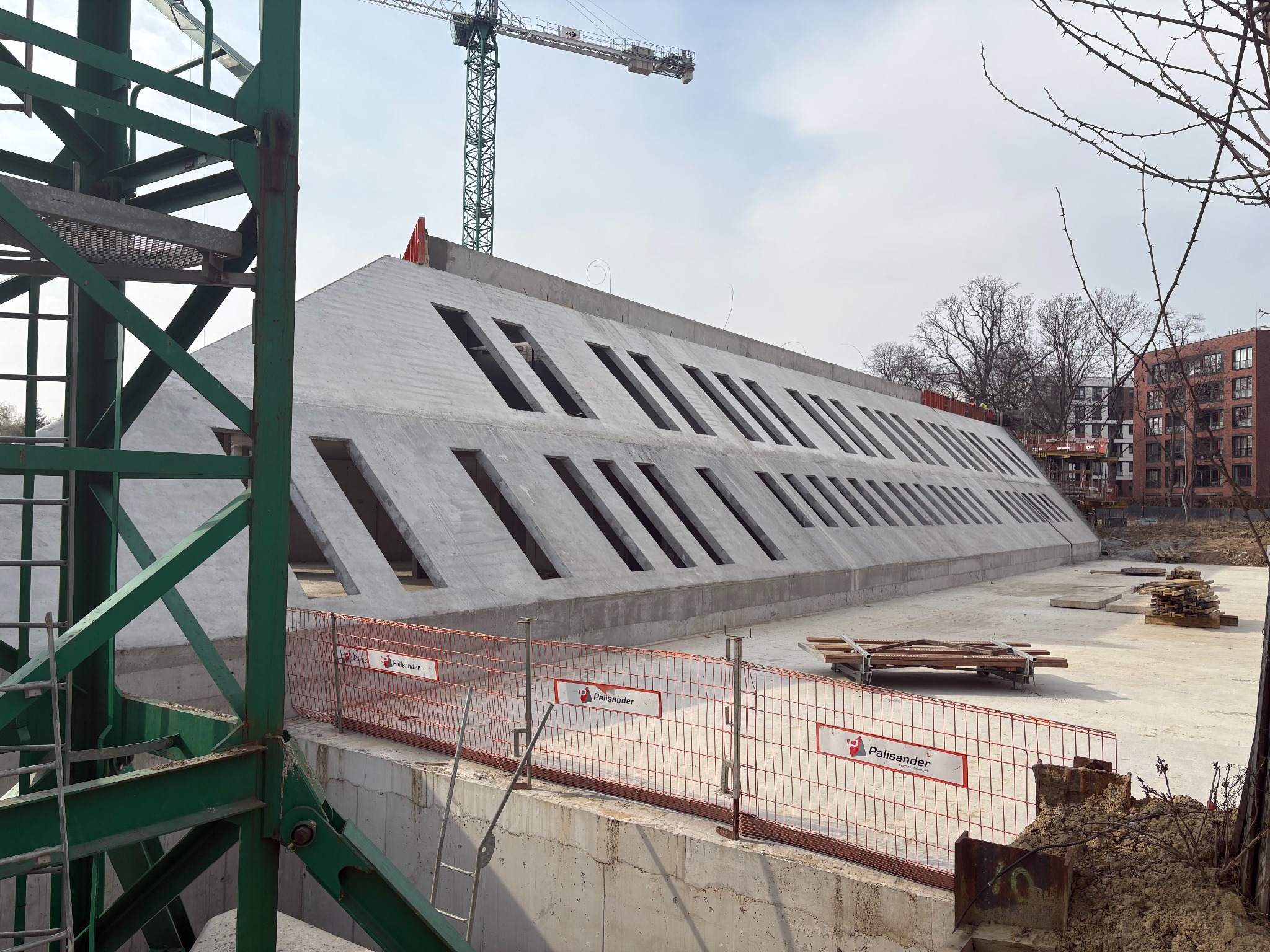 Betonowa konstrukcja budynku z ukośnymi oknami, widoczny żuraw budowlany i ogrodzenie tymczasowe. Surowy stan, plac budowy.