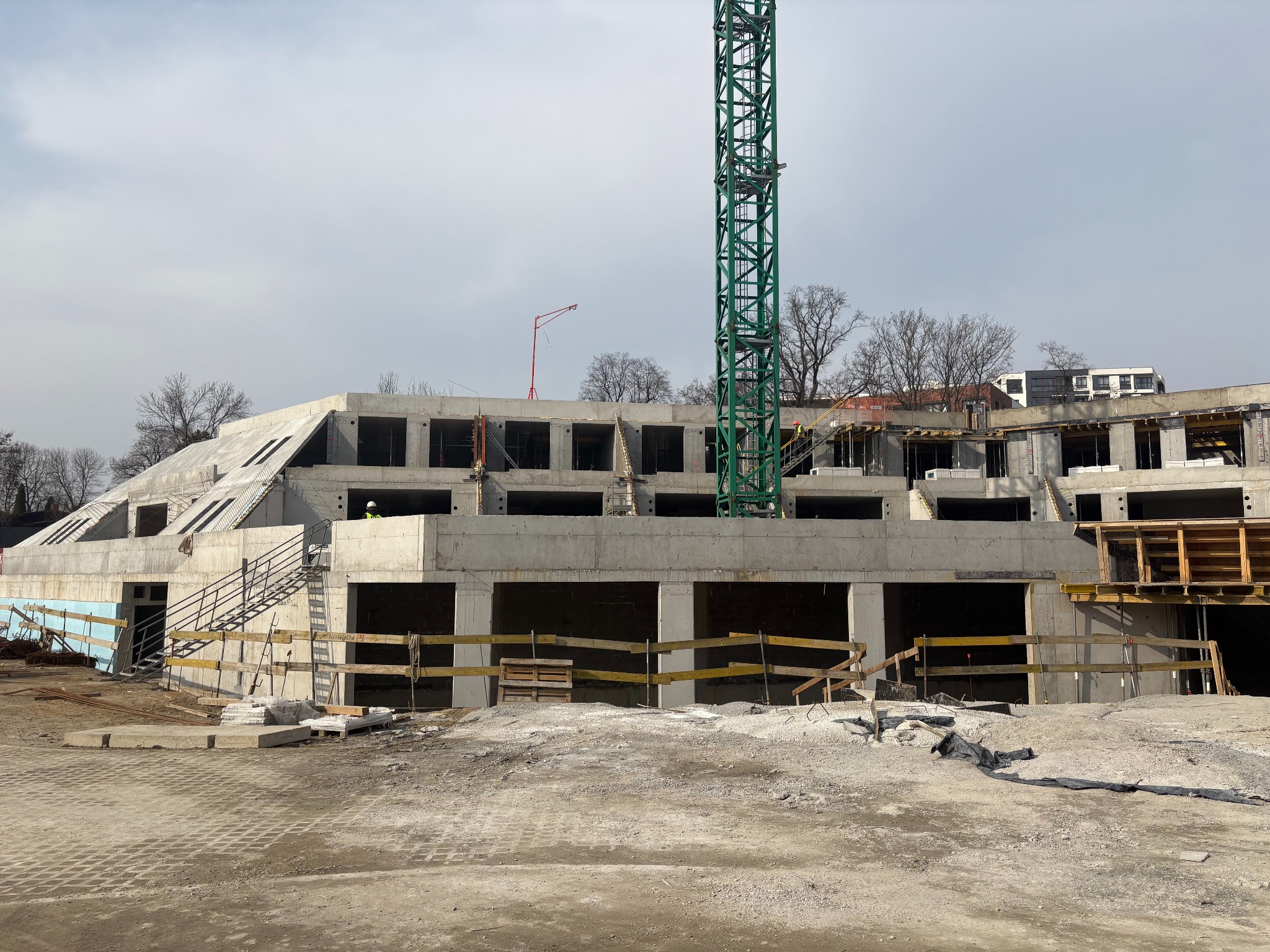 Surowy stan budynku w trakcie budowy z betonu, widoczny żuraw budowlany i szalunki. Budynek wielopiętrowy z otworami okiennymi. Teren budowy.