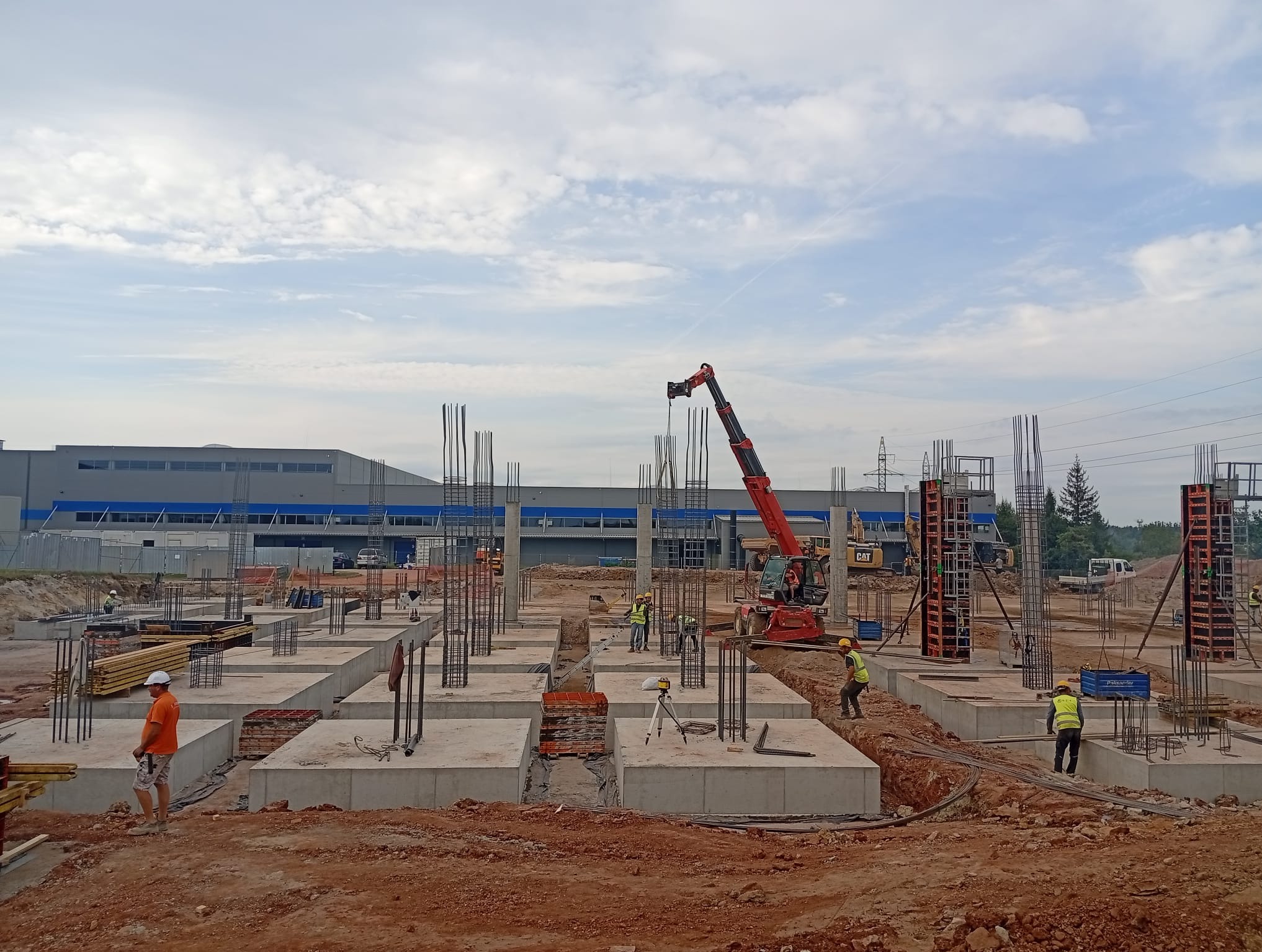 Budowa fundamentów pod halę: betonowe stopy fundamentowe z wystającymi prętami zbrojeniowymi, pracownicy w kamizelkach, dźwig i budynek w tle.