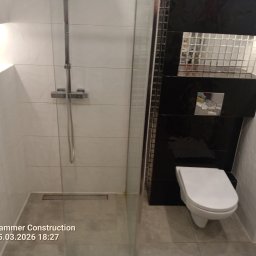 MARIUSZ WÓJCIK Remonty - Nowoczesna łazienka z kabiną prysznicową typu walk-in, białą armaturą i czarnymi akcentami. Ściany wyłożone białymi płytkami, a fragment z toaletą czarnymi, błyszczącymi.