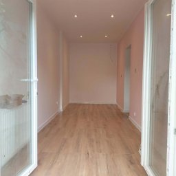 MARIUSZ WÓJCIK Remonty - Jasne panele podłogowe w długim, wąskim pokoju z różowymi ścianami i białymi drzwiami balkonowymi. Widoczne punktowe oświetlenie sufitowe.