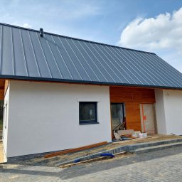 MARIUSZ WÓJCIK Remonty - Nowoczesny dom jednorodzinny z szarą blachą na dachu i drewnianą elewacją, częściowo otynkowany na biało. Widoczne prace wykończeniowe przy wejściu.