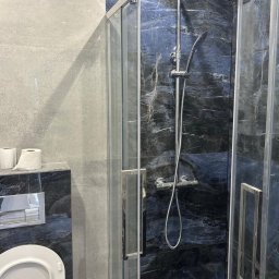 MARIUSZ WÓJCIK Remonty - Wykończona łazienka z kabiną prysznicową typu walk-in, czarne marmurowe płytki z białymi żyłami, szara ściana, toaleta z podwieszanym systemem spłukiwania.