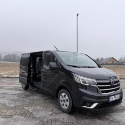 HOME OF LASHES EWELINA OBIREK - Czarny bus Renault Trafic z otwartymi drzwiami przesuwnymi, zaparkowany na kostce brukowej. W tle pole, las i dom. Pochmurny dzień.