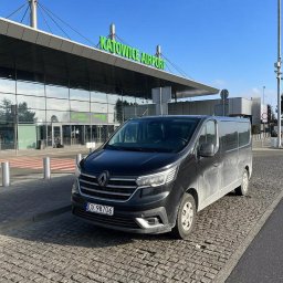 HOME OF LASHES EWELINA OBIREK - Czarny, lekko zabrudzony bus Renault Trafic na kostce brukowej przed budynkiem lotniska Katowice Airport w słoneczny dzień. Tablica rejestracyjna DX 98706.
