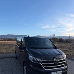 HOME OF LASHES EWELINA OBIREK - Czarny bus Renault Trafic na brukowanym parkingu z widokiem na góry w tle, błękitne niebo. Tablica rejestracyjna DX 98706.