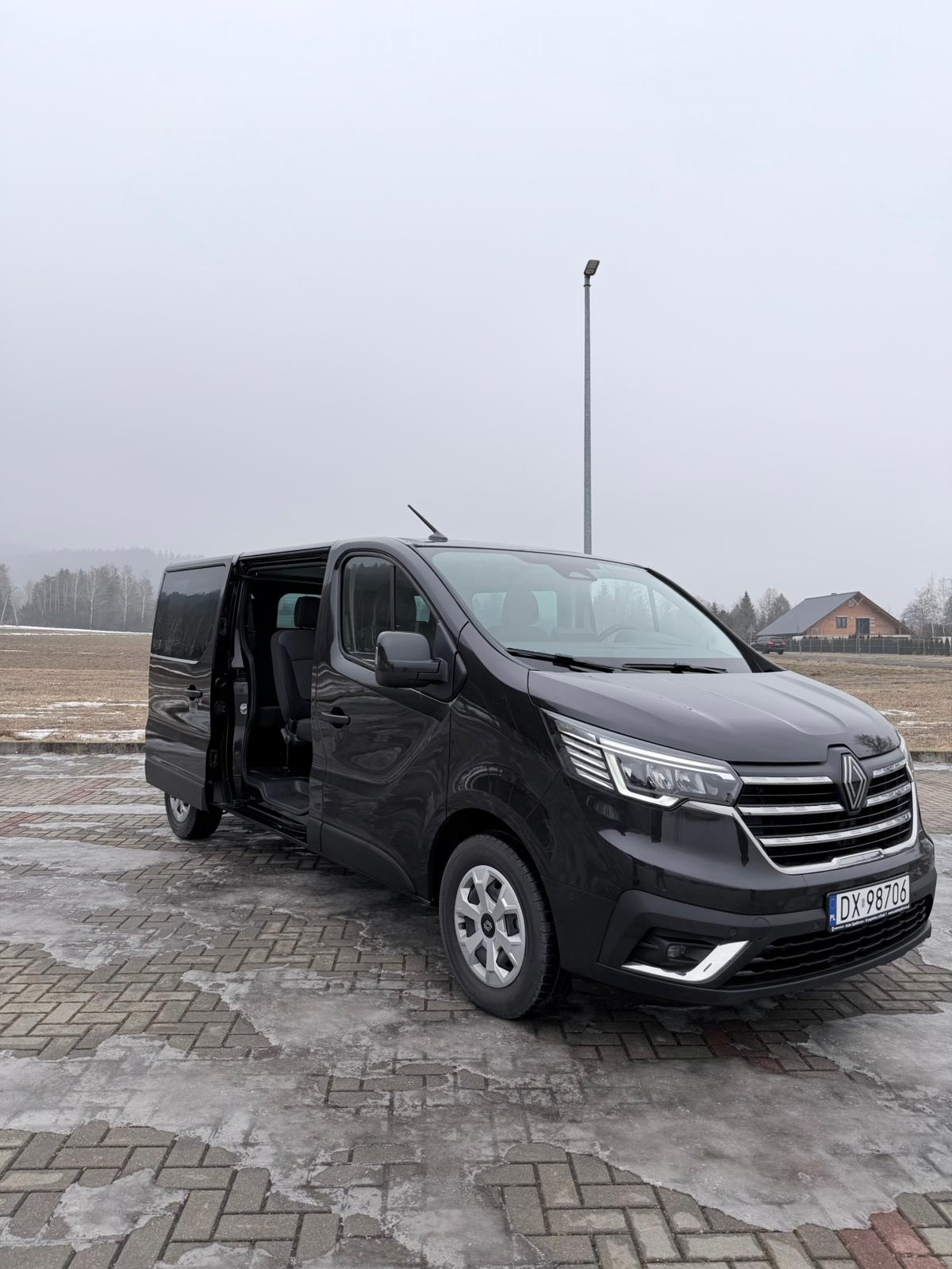 Czarny bus Renault Trafic z otwartymi drzwiami przesuwnymi, zaparkowany na kostce brukowej. W tle pole, las i dom. Pochmurny dzień.