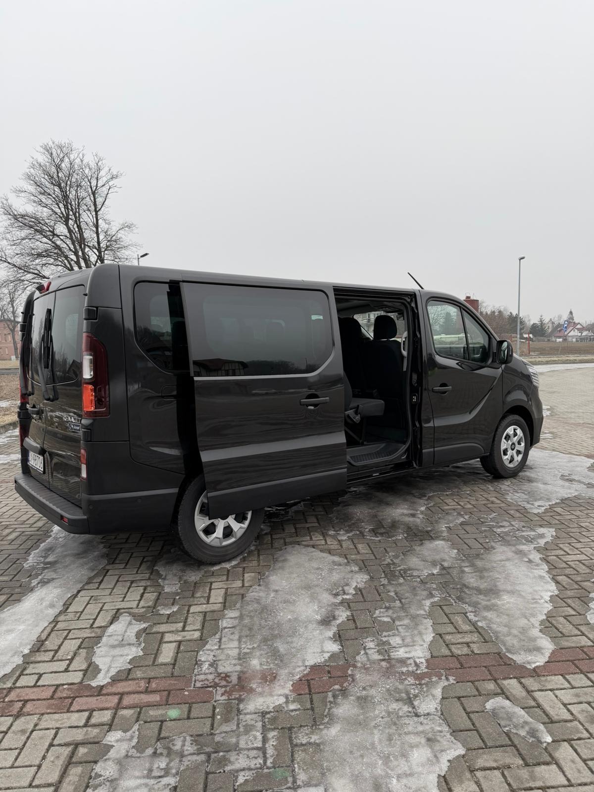 Czarny bus z otwartymi drzwiami przesuwnymi na parkingu z resztkami śniegu. Widoczny fragment wnętrza i siedzenia. Pochmurny dzień.