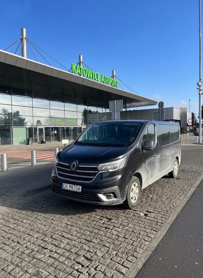Czarny, lekko zabrudzony bus Renault Trafic na kostce brukowej przed budynkiem lotniska Katowice Airport w słoneczny dzień. Tablica rejestracyjna DX 98706.