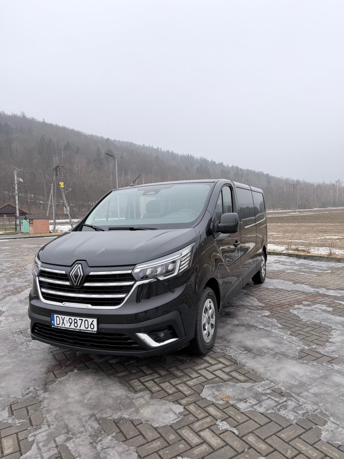 Czarny Renault Trafic z numerem rejestracyjnym DX 98706, zaparkowany na kostce brukowej, z zalesionym wzgórzem w tle, w pochmurny dzień.