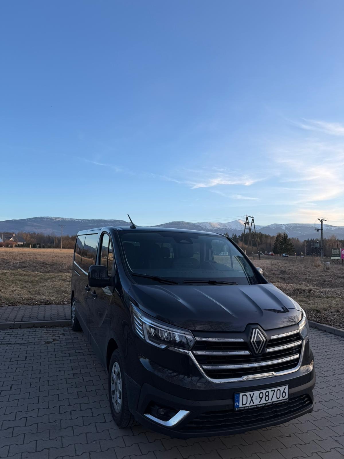 Czarny bus Renault Trafic na brukowanym parkingu z widokiem na góry w tle, błękitne niebo. Tablica rejestracyjna DX 98706.