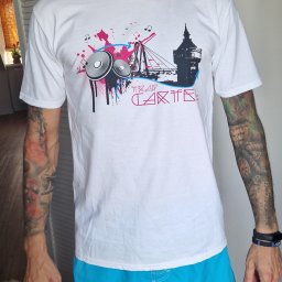 EMBE Maciej Piotrowski - Biała koszulka z unikalnym nadrukiem: wieża, głośniki, nuty i napis 'Trap Cartel'. Mężczyzna w niebieskich szortach prezentuje koszulkę. Widoczne tatuaże na ramionach.